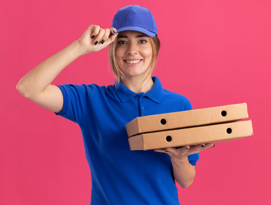 披萨身着制服 面带微笑的年轻漂亮送货女孩把手放在帽子上 手里拿着粉色的比萨饼盒姿势帽子漂亮