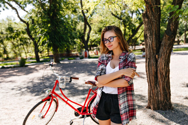 骑车人优雅的金发女士戴着眼镜在骑自行车后摆姿势穿着红色自行车的温文尔雅女孩的户外肖像健身成人休闲