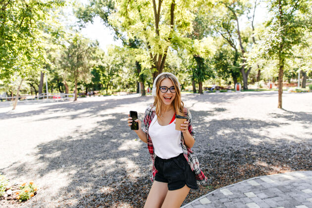 卷发优雅的金发女士 穿着黑色短裤 在公园里欢笑美轮美奂的欧洲女孩 在夏天的周末 拿着一杯咖啡到处闲逛公园阳光微笑