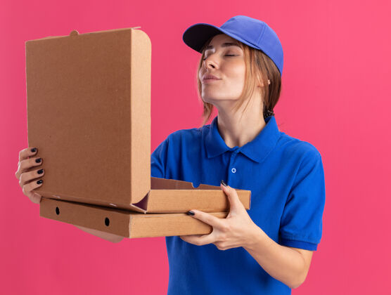 盒子穿着制服的年轻漂亮的送货女孩拿着比萨饼盒 假装嗅着粉红色的味道漂亮送货制服