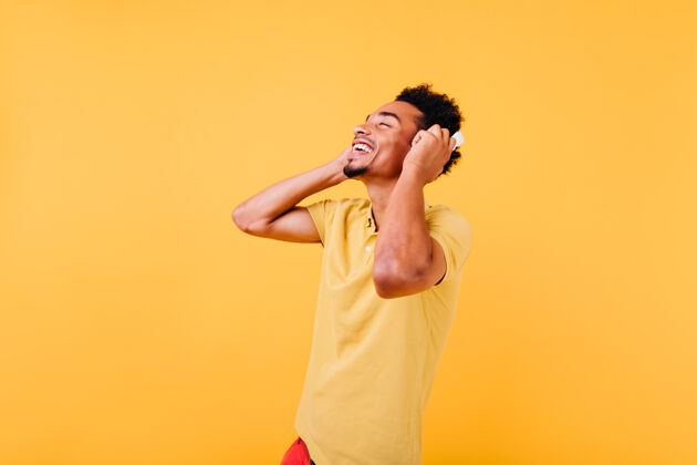 黄色幸福的男人 带着美丽的微笑 听着音乐非洲男模戴着耳机在室内拍摄 表达积极的情绪情感非洲学生