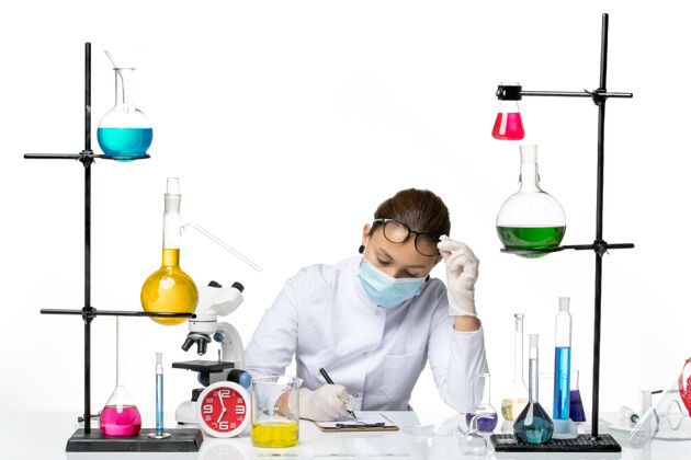 实验室前视图穿着医疗服的女化学家 戴着面罩 坐着 拿着溶液 在白色背景上写笔记 溅起实验室病毒化学冠状病毒病毒解决方案坐