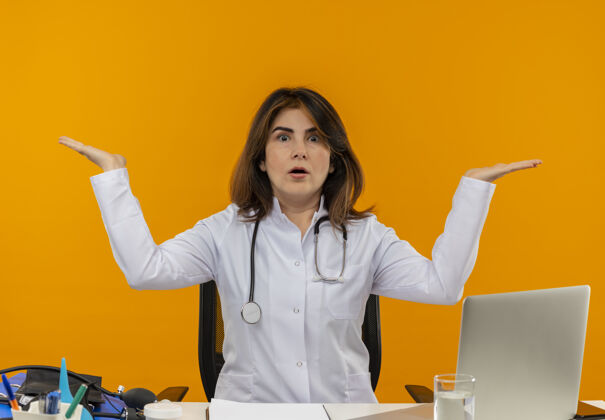 空令人印象深刻的中年女医生穿着医用长袍和听诊器坐在办公桌前 医疗工具剪贴板和笔记本电脑显示空手孤立手中年女性