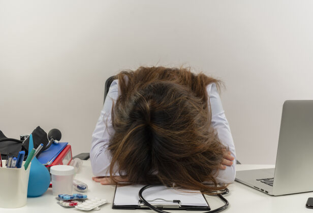 医疗疲惫的中年女医生穿着医用长袍和听诊器坐在办公桌旁 用医疗工具剪贴板和笔记本电脑把头靠在办公桌上穿医生听诊器