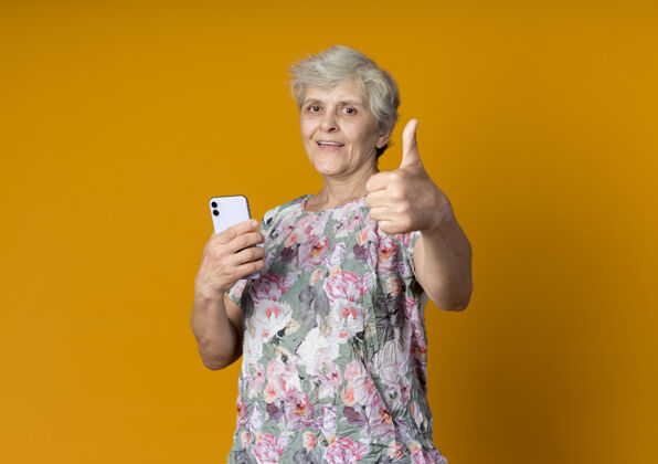 拇指快乐的老妇人拿着电话 竖起大拇指孤立地站在橙色的墙上电话抱着向上