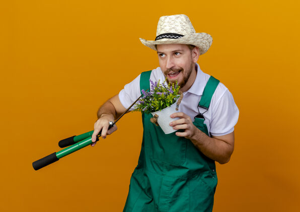 人高兴的年轻英俊的斯拉夫园丁穿着制服 戴着帽子 手持花草和修剪者抬头孤立地看着抱着男人花园