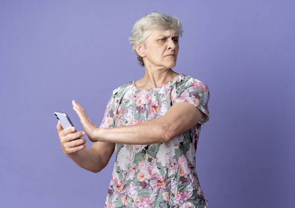 女人不高兴的老妇人拿着电话假装推着看紫色墙上孤立的一面推抱紫色