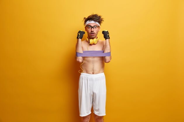 眼镜运动员用健身口香糖锻炼手部肌肉 用震惊的表情凝视 穿白色短裤 戴头带 使用立体声耳机 隔离在黄色墙壁上保持强壮和健康设备运动长度