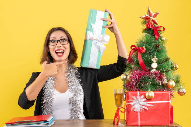 礼物一位身着西装 面带微笑的商务女士 戴着眼镜 惊讶地指着自己的礼物 坐在一张桌子旁 桌子上放着一棵圣诞树树惊喜成人