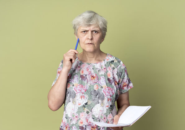橄榄困惑的老妇人拿着笔记本 把笔孤立地放在橄榄绿的墙上老拿着钢笔