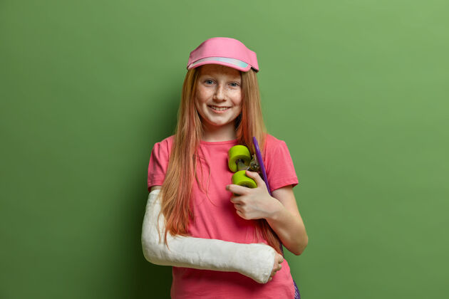 手术笑容可掬的红发女孩在玩滑板后发生意外 手臂骨折后戴石膏 保持快乐 在喜爱的运动中受伤 靠着绿墙站着儿童 保健外观情绪创伤