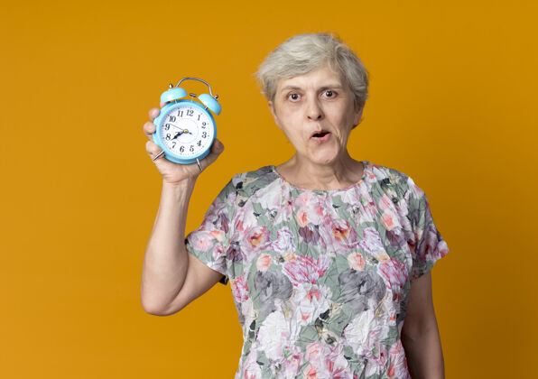 老人兴奋的老妇人把闹钟隔离在橙色的墙上闹钟女人兴奋