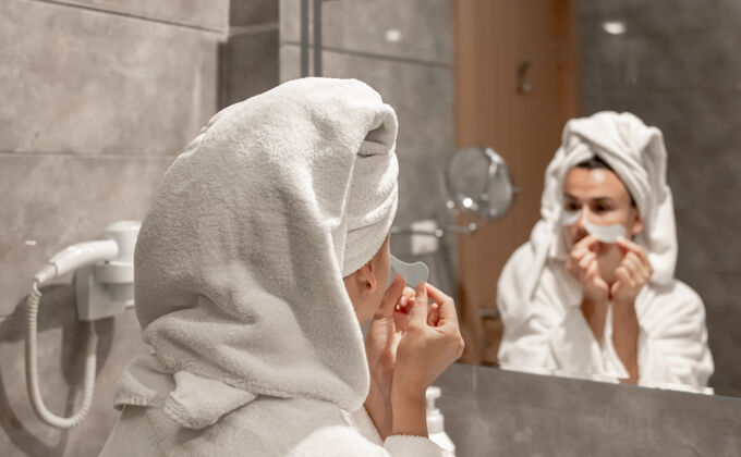 浴袍一个女孩穿着浴衣 头上戴着毛巾 在镜子前的浴室里 把补丁贴在眼睛下面毛巾水疗可爱