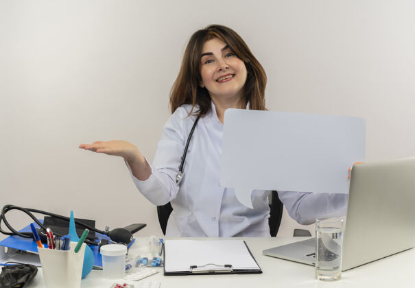 人微笑的中年女医生穿着医用长袍和听诊器坐在办公桌旁 拿着医疗工具剪贴板和笔记本电脑 拿着聊天泡泡 显示空手孤立空坐长袍