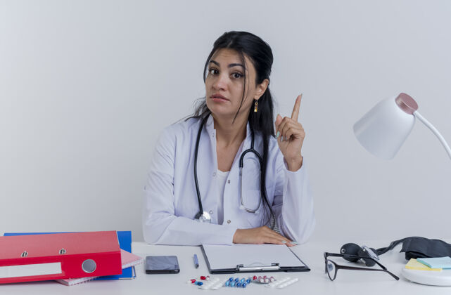 医生令人印象深刻的年轻女医生穿着医用长袍和听诊器坐在办公桌旁 手持医疗工具 手放在办公桌上抬起手指提高女人印象