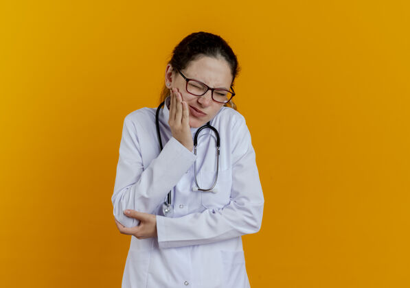 女性闭着眼睛的年轻女医生穿着医用长袍 戴着眼镜的听诊器把手放在疼痛的牙齿上医生长袍眼睛
