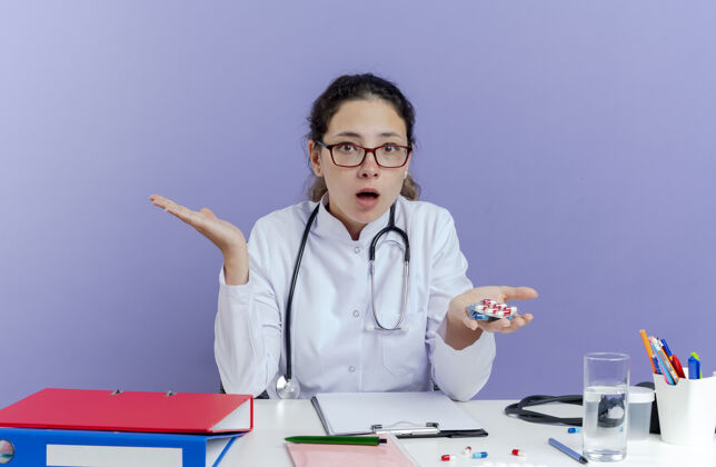 抱着令人印象深刻的年轻女医生穿着医用长袍和听诊器坐在办公桌旁 手里拿着医疗工具 手里拿着药片 空手看上去很孤立手人工具