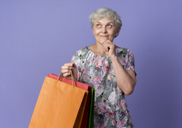 购物高兴的老妇人把手放在下巴上 手里拿着纸购物袋 孤零零地仰望紫色的墙上女人手拿着