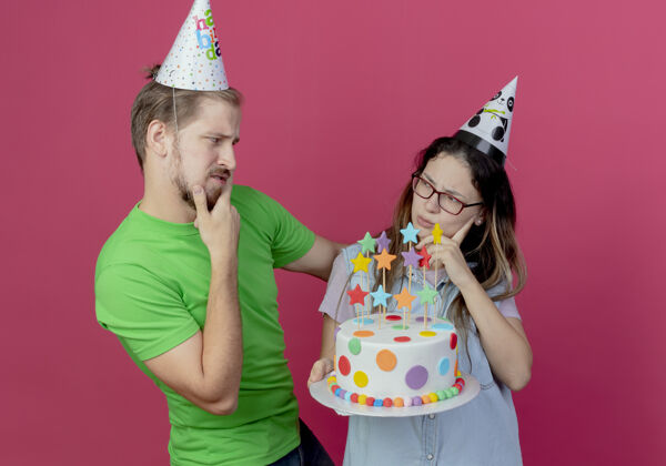 拿着困惑的年轻夫妇戴着派对帽看着对方 生日女孩拿着蛋糕隔离在粉红色的墙上困惑每人帽子