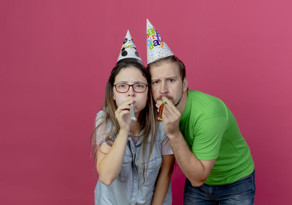 粉色一对戴着派对帽的年轻夫妇不高兴地看着彼此 在粉红色的墙上孤立地吹着口哨每个生日吹口哨