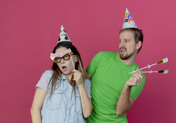 香槟不高兴的年轻人戴着派对帽拿着假香槟酒杯在棍子上看着惊讶的年轻女孩拿着眼罩在棍子上隔离在粉红色的墙上假的派对生日