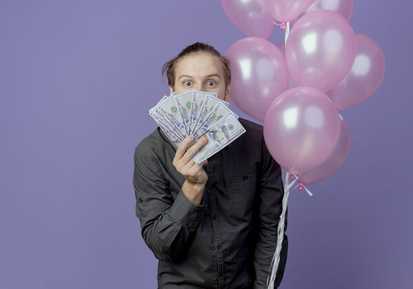 站着兴奋的帅哥手持氦气球站在紫色的墙上 看着孤立的金钱生日气球紫色