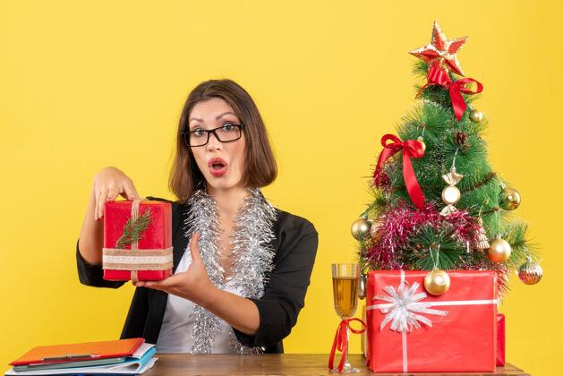 桌子一位穿着西装 戴着眼镜 拿着礼物 坐在一张桌子旁 桌上放着一棵圣诞树的好奇而惊讶的商务女士快乐人大人