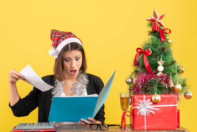 文件惊艳迷人的女士穿着西装 戴着圣诞老人的帽子和新年的装饰品 手里拿着文件 在办公室里的黄色隔离圣诞老人圣诞老人年份