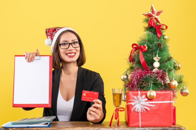 微笑微笑迷人的女士穿着西装 戴着圣诞老人的帽子和眼镜 在黄色的办公室里展示着银行卡和文件人表演成人