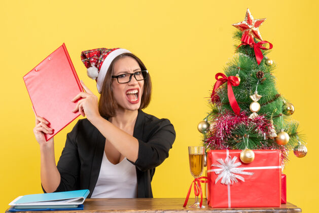 肖像情绪紧张的迷人女士西装革履戴着圣诞老人的帽子和眼镜在黄色的办公室里与世隔绝圣诞老人快乐情绪
