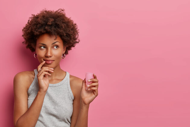 干燥沉思的非洲裔美国妇女持有有效安全实惠的月经产品 很好的防漏 选择可重复使用的月经杯 思考如何使用它 粉色墙上的空白处渗漏民族替代
