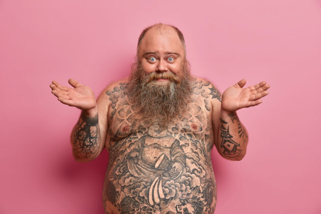脂肪犹豫不决的开朗男人的照片摊开手掌 说我不知道 高兴和困惑 有大肚子 纹身 不知道如何适合和松散的重量 隔离在粉红色的墙上麻烦积极成人