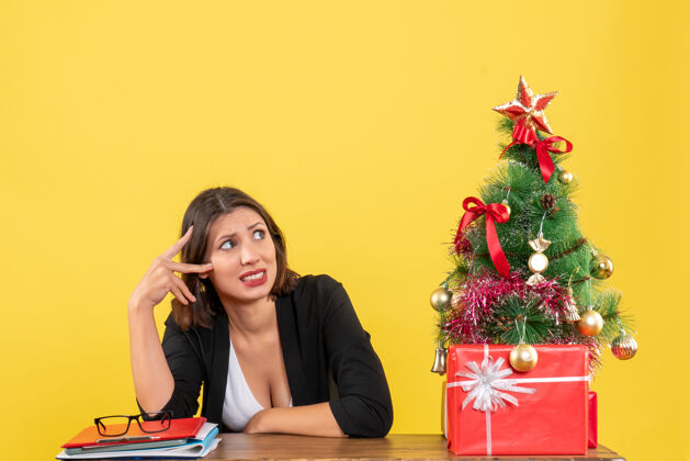 漂亮这位年轻漂亮的女士惊奇地坐在黄色办公室装饰的圣诞树旁的桌子旁 对什么东西感到困惑快乐人靠近