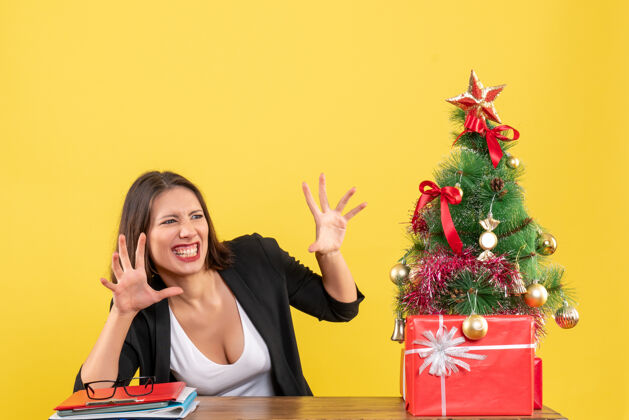 年轻女人一个有趣的年轻女子坐在黄色办公室装饰的圣诞树旁的桌子旁 带着惊讶的表情看着什么东西成人搞笑快乐