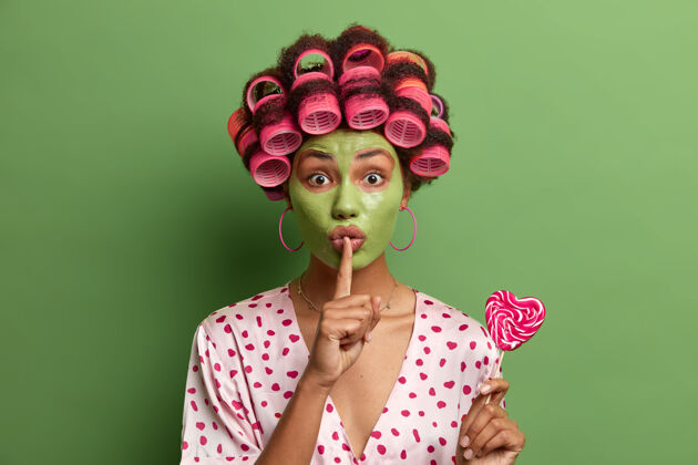 滚轴神秘的少数民族妇女戴着绿色的面具和卷发器 默默地做手势 告诉秘密 拿着美味的心形棒棒糖 做发型 穿礼服 在家里花时间应用棒棒糖卷发