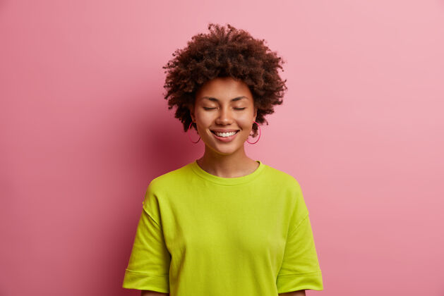 非洲美丽开朗的女人的肖像闭上眼睛 愉快地微笑 穿着休闲的绿色t恤 听到愉快的支持的话 孤立在粉红色的墙壁快乐的情绪和感情立场姿势情绪