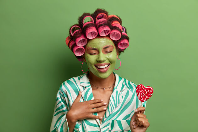 卷发喜气洋洋的美女用卷发器和绿色的面膜 开心地笑着 在家里享受美容过程中的乐趣 把棒棒糖放在手杖上 准备派对皮肤呵护 健康 SPA身体新鲜适用