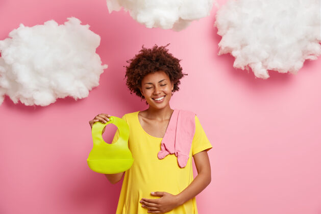 期望正喜的女人期待着新生的孩子 呵护着肚子 抱着围嘴和婴儿的衣服 在回忆她怀孕的时候拍照期待和母爱的概念微笑女人怀孕