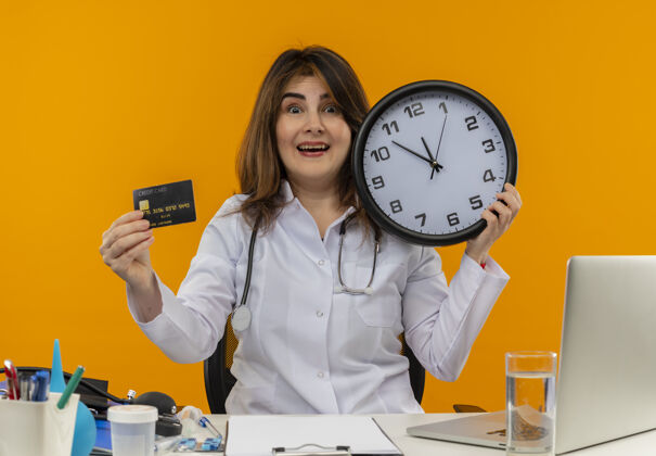 医疗令人印象深刻的中年女医生穿着医用长袍和听诊器坐在办公桌前 医疗工具剪贴板和笔记本电脑拿着时钟和信用卡隔离持有穿着橙色