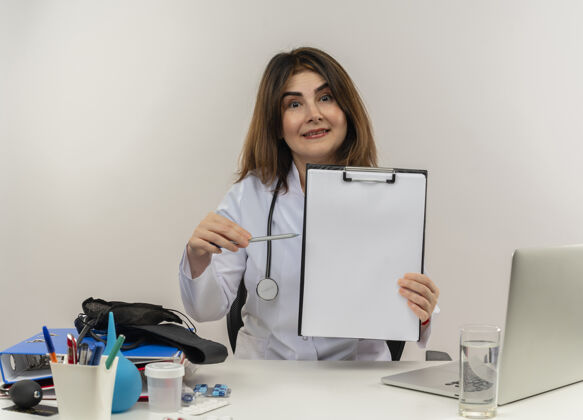工具印象深刻的中年女医生穿着医用长袍和听诊器坐在办公桌旁 手持医疗工具和笔记本电脑 用钢笔指着剪贴板指着桌子成人