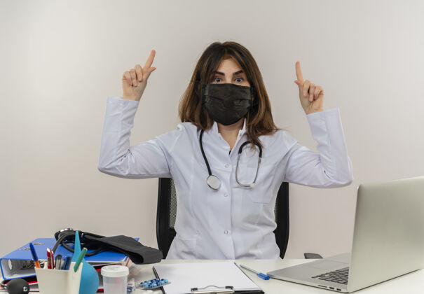 剪贴板印象深刻的中年女医生戴着医用口罩坐在办公桌前 用医疗工具剪贴板和笔记本电脑指着隔离人医疗面具