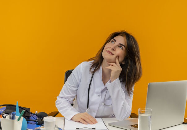坐着高兴的中年女医生穿着医用长袍和听诊器坐在办公桌前 用医疗工具剪贴板和笔记本电脑触摸脸 抬头看去笔记本电脑剪贴板工具