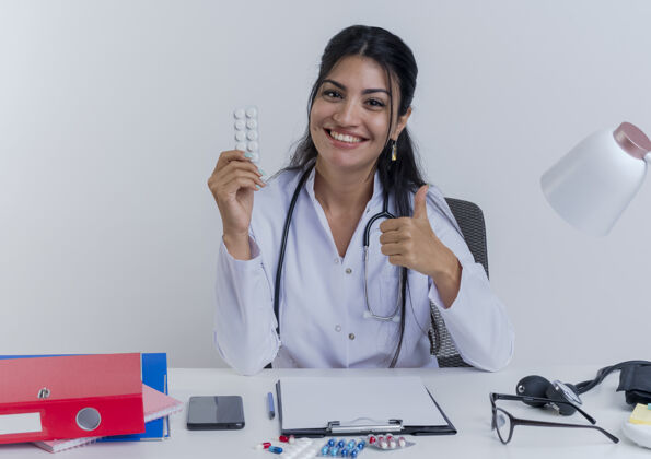 医疗快乐的年轻女医生穿着医用长袍和听诊器坐在办公桌旁 手里拿着医疗工具 手里拿着一包药片 神情孤立地竖起大拇指向上表演成人