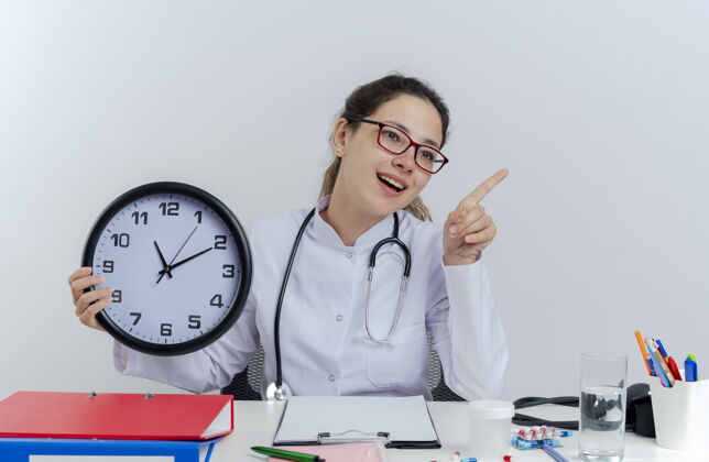 眼镜年轻的女医生穿着医用长袍 戴着听诊器 戴着眼镜 坐在办公桌旁 手里拿着医疗工具 拿着时钟 望着被隔离的一边指尖女性印象