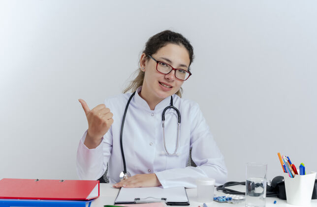 听诊器年轻的女医生穿着医用长袍 戴着听诊器 戴着眼镜 坐在办公桌旁 手里拿着医疗工具 脸上露出孤立的拇指请桌子表演