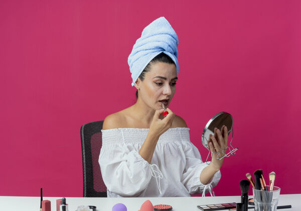 应用自信漂亮的女孩裹着发巾坐在桌子上 拿着化妆工具拿着唇膏 看着镜子挂在粉红墙上坐漂亮工具