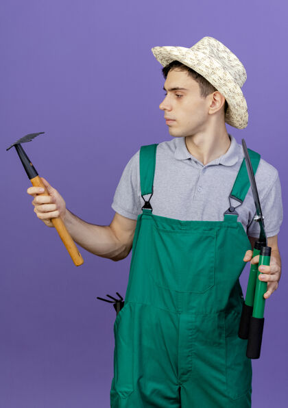 复制自信的年轻男园丁戴着园艺帽站着 拿着园艺工具 看着紫色背景上孤立的耙子 还有复制空间男性空间帽子