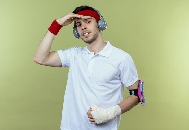 耳机戴着头戴式耳机和智能手机臂带的年轻运动型男子远远望去 手举着头站在绿色背景上年轻头头带
