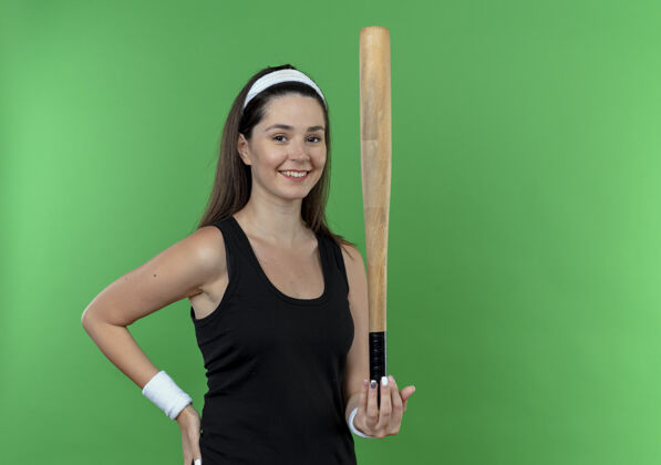 健身戴着头巾的年轻健身女士手持棒球棒 站在绿色背景下 面带微笑地看着镜头站着年轻脸