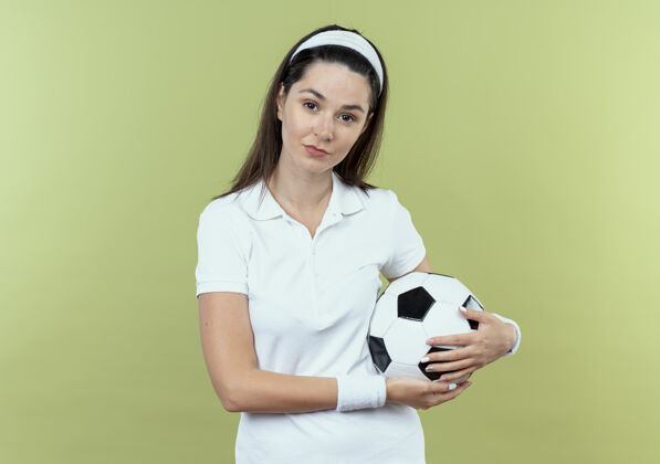 自信戴着头巾拿着足球的年轻健身女士站在灯光背景下 自信地看着摄像机相机健身头带